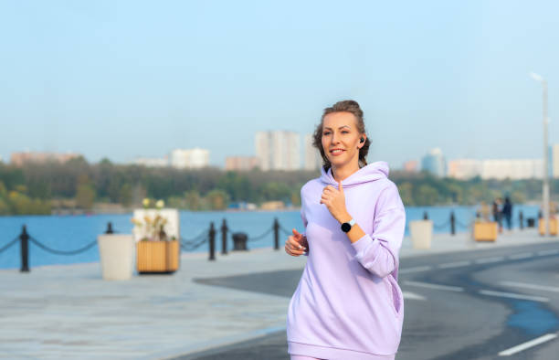 Young woman running on the city road. The girl runs along the city embankment and listens to music. An active life position, the pleasure of a morning run and a good body condition. medium shot stock pictures, royalty-free photos & images