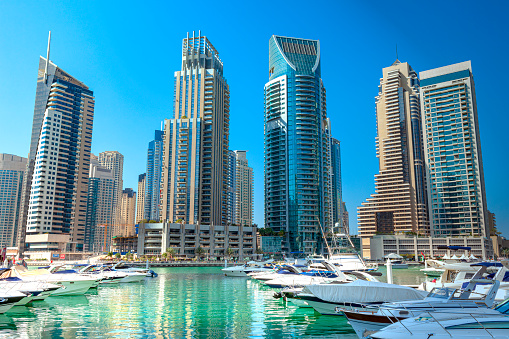 Dubai, United Arab Emirates - In view are boats and motor yachts moored on Dubai Marina; in the background are modern skyscrapers that house offices, apartments and hotels. Photo shows off a modern luxurious lifestyle that is synonymous with the Arabian Gulf city of Dubai. Photo shot in the afternoon sunlight against a clear blue sky; horizontal format. Note to Inspector: All logos from boats and registration numbers have been removed. In the far background, there are more than three logos, but there is not much difference between them in size.