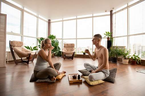 Young restful man and woman with crossed legs sitting on pillows in front of one another and having herbal tea after yoga training at home