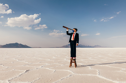 A young teenager businessman dressed in a suit stands on the Bonneville Salt Flats of Utah, USA, looking for business opportunities through the COVID-19 pandemic. He is wearing a face mask and looking through a telescope searching for business profitability and success.