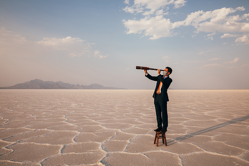 A businessman dressed in a suit stands on the Bonneville Salt Flats of Utah, USA, looking for business opportunities through the COVID-19 pandemic. He is wearing a face mask and looking through a telescope searching for business profitability and success.
