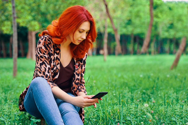 a mulher está mandando sms enquanto está sentada na grama. - mobilestock freedom enjoyment blue - fotografias e filmes do acervo