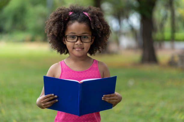 afroamerikanische kleine mädchen mit lockigen und haare tragen brille beim lesen eines buches im sommer im freien. bildungs- oder exkursionskonzept - child glasses eyewear little girls stock-fotos und bilder