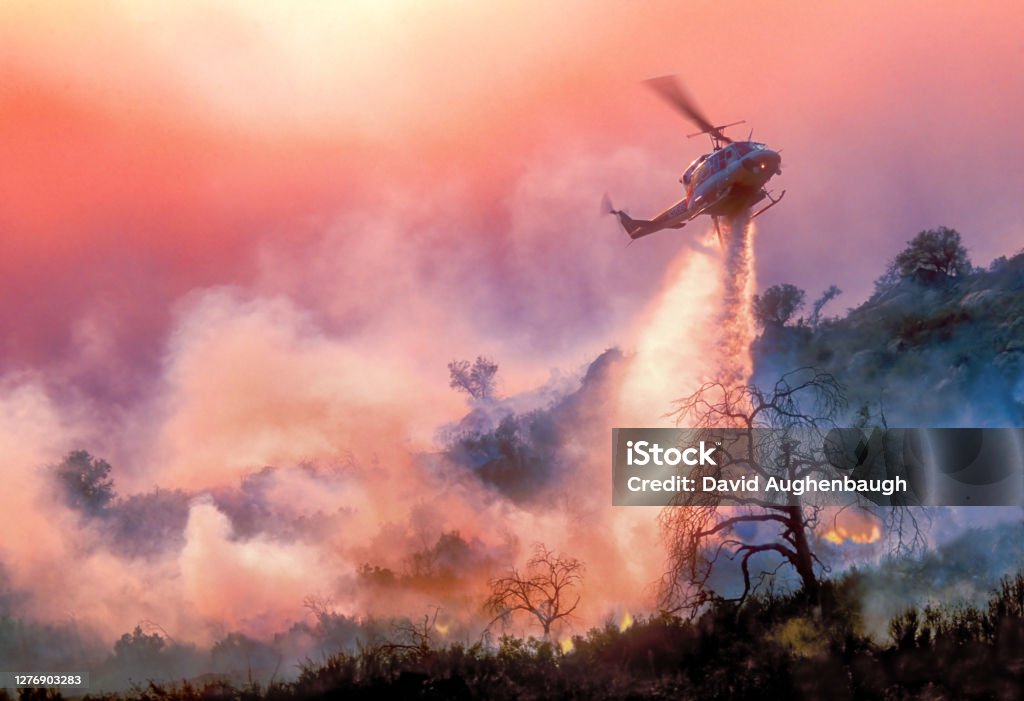 Helicopter Water-Drop on California Wildfire A helicopter dropping water on a California wildfire in rugged terrain, backlit by a setting sun filtered through multiple layers of smoke Climate Change Stock Photo
