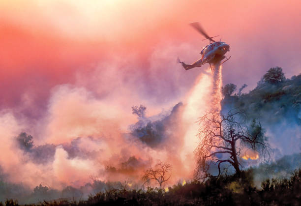 Helicopter Water-Drop on California Wildfire A helicopter dropping water on a California wildfire in rugged terrain, backlit by a setting sun filtered through multiple layers of smoke firefighter photos stock pictures, royalty-free photos & images