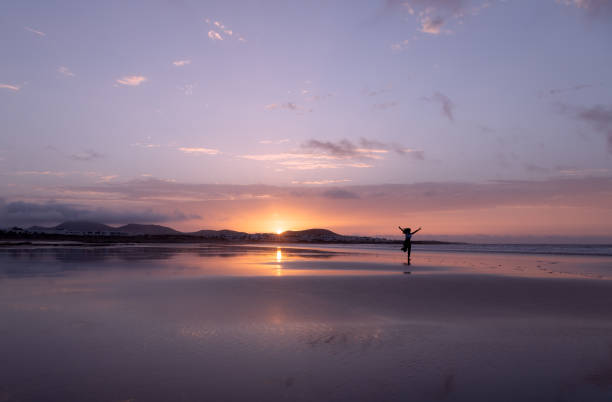 sylwetka nierozpoznawalnej kobiety, biegającego z otwartymi ramionami na plaży famara o zachodzie słońca na lanzarote, wyspy kanaryjskie, hiszpania. - famara zdjęcia i obrazy z banku zdjęć