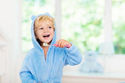 Child brushing teeth. Kids tooth brush and paste. Little baby boy in blue bath robe or towel brushing his teeth in white bathroom with window on sunny morning. Dental hygiene and heath for children.