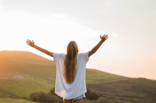 Young girl with a white t-shirt with her two arms raised as she is enjoying her freedom in nature. She is standing in a mountain hill during the golden hour.