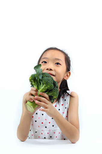 Portrait of cute little Asian girl holding a broccoli, sitting and looking up againts white background