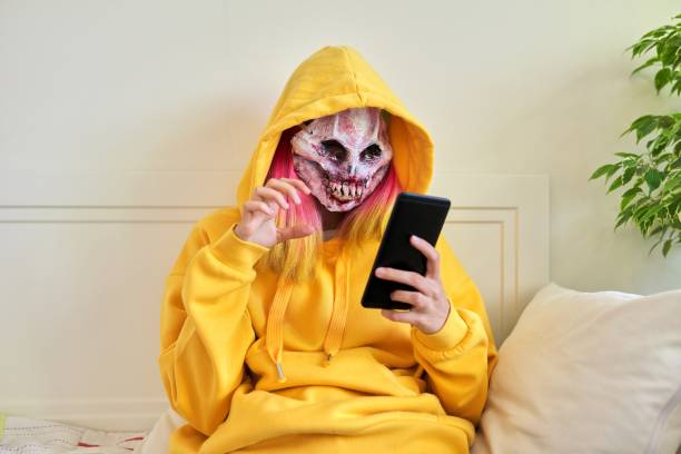 halloween party at home, teenage girl in scary hoodie mask with smartphone - face paint human face mask carnival imagens e fotografias de stock