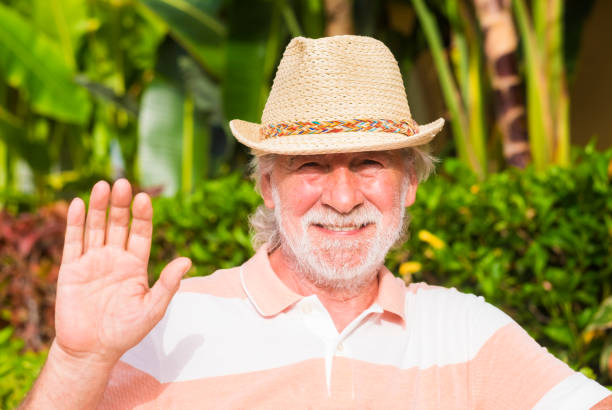 istock Attractive bearded senior man with straw hat greets with one hand looking at the camera while standing against a stone wall - concept of active seniors during retirement 1276921855