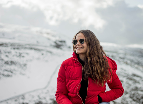Young dark-haired woman with sunglasses at the top of the mountain with snow smiling and happy. Cold weather. Sitting on a rock with a beautiful landscape with snow and big mountains behind.