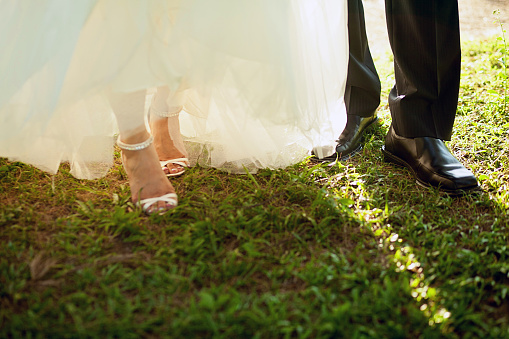 Low-perspective of groom's black shoes and bride's white high hills under whose wedding dress. Bride and groom walking on the grass field.