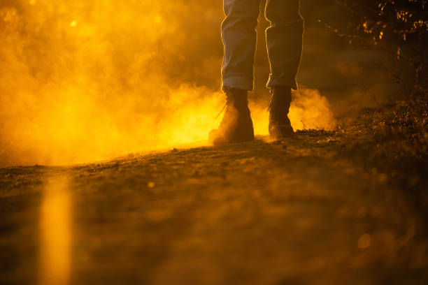 A traveler in boots walks along a dusty road at sunset.