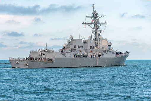 One crew member is raising two maritime flags, while the other watches or assists, on the bridge of USS Canberra (LCS 30), an Independence Class combat ship of the US Navy which is docked at Garden Island in Sydney Harbour.  She was in port for her commissioning ceremony on 22 July 2023.  As part of the ceremony, her captain was presented with a kangaroo insignia attached to all ships of the Royal Australian Navy, but decorated in the colours and design of the Stars and Stripes.  That insignia has been attached to the ship and visible on the right.  The name of the ship and serial number is visible on the orange life ring.  A large, padded chair is visible on the bridge.  This image was taken on a cloudy afternoon on 29 July 2023.