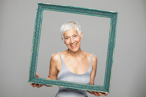 Portrait of beautiful mature woman with grey hair looking at camera. She is holding  green picture frame and looking through it. Attractive middle aged woman with beautiful smile isolated over grey background.