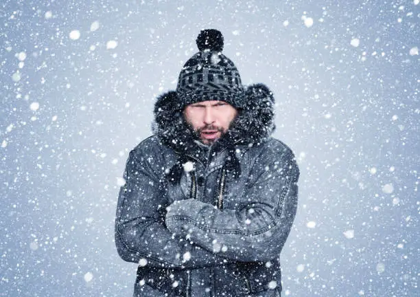 Photo of Frozen bearded man in winter clothes warms his hands, cold, snow, frost, blizzard