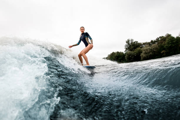 istock Smiling woman in wetsuit rides down on surf style wakeboard on wave 1276991590
