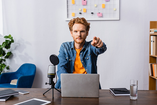 serious radio host in denim shirt pointing with finger at camera while sitting at workplace