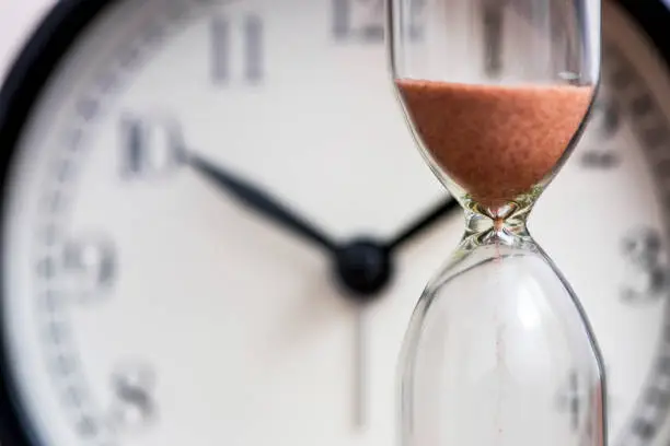 Photo of Hourglass on the background of office watch as time passing concept for business deadline, urgency and running out of time. Sand clock, business time management concept