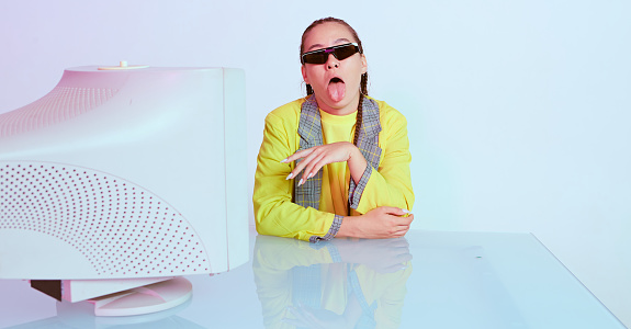 Stylish asian female office worker in sunglasses sitting with obsolete computer and shows tongue. Colored Neon light. High quality photo