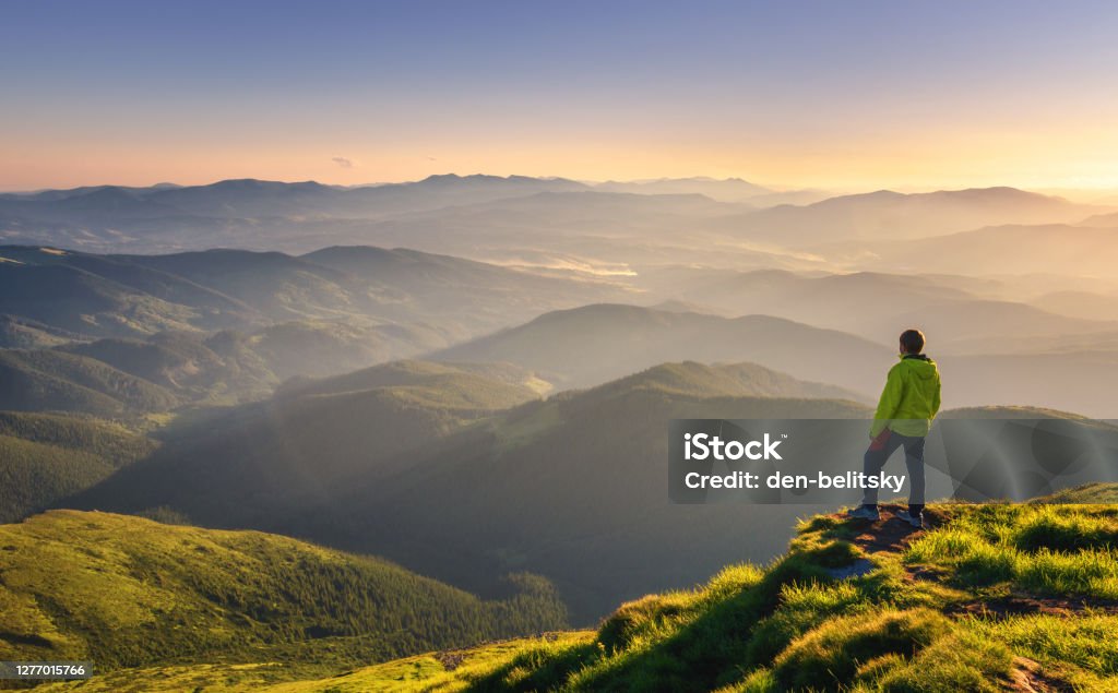 Sporty man on the mountain peak looking on mountain valley with sunbeams at colorful sunset in autumn in Europe. Landscape with traveler, foggy hills, forest in fall, amazing sky and sunlight in fall Mountain Stock Photo