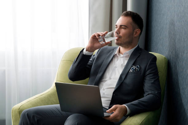 istock Young businessman is using laptop and drinking water while sitting at the hotel room with suitcase 1277015947