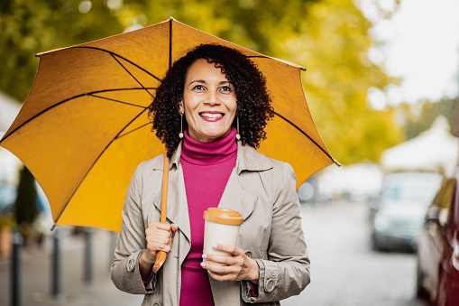 Photo of mature woman with umbrella outdoors smiling and holding reusable coffee cup