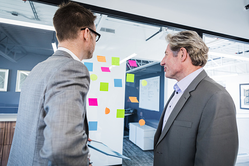 Two male colleagues at a modern office, standing in front of a glass wall with colorful sticky notes on it. Profile view.