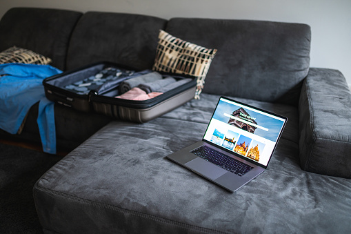 Laptop with a travel website open and a luggage case lying on couch