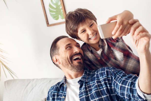 istock Father and little son at home sitting on sofa taking selfie on smartphone looking camera grimacing playful close-up 1277041475