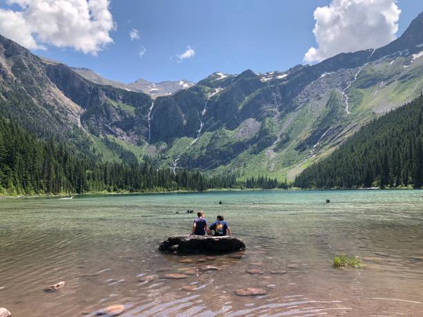 Two children on a log in the middle of lake enjoying the beauty of Avalanche Lake Glacier National Park with many waterfalls in the distant