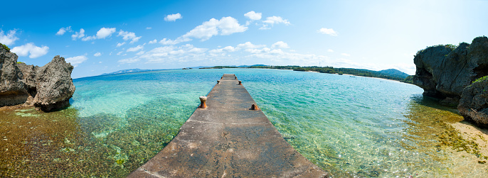A fisheye panorama showing a concrete fishing jetty or pier in Onna village, Okinawa jutting out into the clear blue ocean. This area is popular for resorts and marine activities.