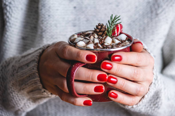 A woman in a warm cozy sweater is holding a cup of hot chocolate decorated with marshmallows, red candy and a Christmas tree branch. Concept of a festive mood and enjoying a hot beverage A woman in a warm cozy sweater is holding a cup of hot chocolate decorated with marshmallows, red candy and a Christmas tree branch. Concept of a festive mood and enjoying a hot beverage. red nail polish stock pictures, royalty-free photos & images