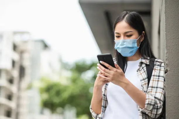 Photo of Young Asian beautiful woman travelers use their mobile phones to find travel locations and call for taxi transportation. She wearing protection mask for new normal safety travel trips.