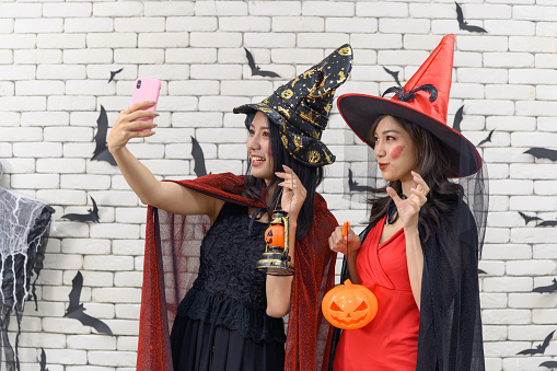 Portrait of two beautiful young asian woman wearing witch costume holding pumpkin Jack O Lantern taking selfie photo on mobile phone
