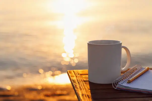 mug of coffee and notebook on table with sea at sunrise on the background
