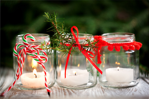 Christmas decorations. Candles in glass jars with fir on holiday background. Cozy handmade holiday home decor: glass jar with candle decorated with red ribbon.