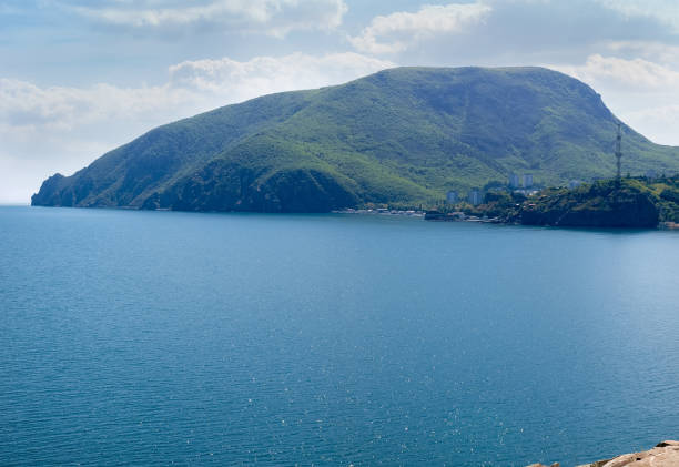 Coastal mountain of dome-like form overgrown with forest near the sea with water surface on a foreground. Mount Ayu-Dag, Crimea. View from Cape Plaka.