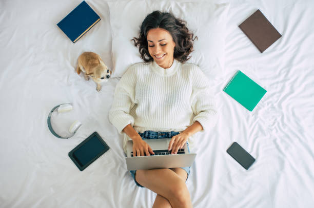 fofa, a jovem e linda está deitada na cama em casa com seu cachorrinho e algumas coisas para estudar. garota com laptop e telefone - dog school audio - fotografias e filmes do acervo