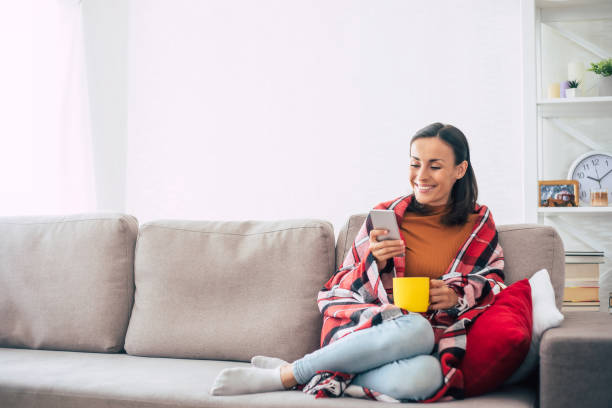 feliz hermosa joven con un teléfono inteligente y una taza en las manos está cubierto bajo una trenza a cuadros en el sofá mientras descansa en casa. temporada de invierno y vacaciones de navidad - living room people joy happiness fotografías e imágenes de stock