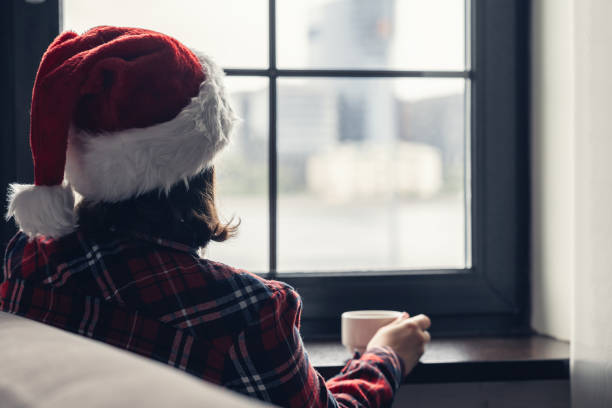 achtermening van jonge vrouw in een rode kersthoedzitting van de kerstman in de buurt van het raam, ontbijten met een kopje koffie. concept - eenzaam stockfoto's en -beelden