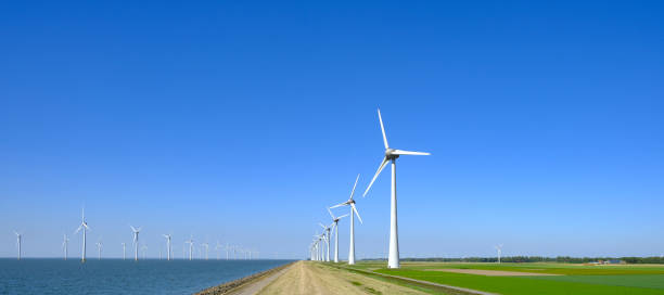 Wind turbines in a windpark on the shore of a lake in The Netherlands Row of wind turbines in a waindpark next to a levee in the IJsselmeer in Flevoland, The Netherlands, with a blue sky above. flevoland photos stock pictures, royalty-free photos & images