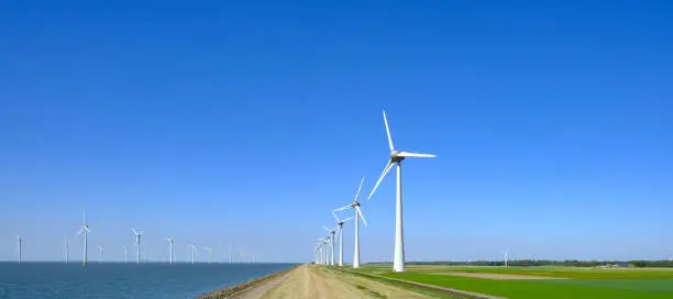 Row of wind turbines in a waindpark next to a levee in the IJsselmeer in Flevoland, The Netherlands, with a blue sky above.