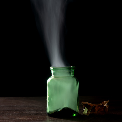 Photo of green poison bottle with smoke in dark. No people are seen in frame. The background is black. Shot with a full frame mirrorless camera.