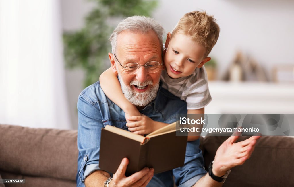 . Cheerful grandfather and grandson reading book together Happy family elderly  man and little boy smiling r while sitting on couch and reading fascinating fairy tale together at home Grandfather Stock Photo