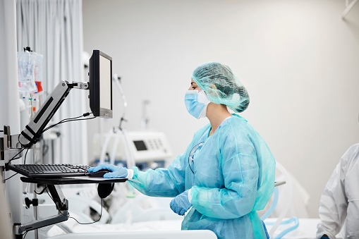 Side view of nurse using computer in ward. Female healthcare worker is working in hospital during coronavirus outbreak. She is wearing protective workwear.