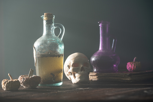 Three rotten pumpkins, a black antique book and a bottle full of dirty poisonous liquid on wooden desk for halloween concept. The background is foggy and black. Shot with a full frame mirrorless camera.