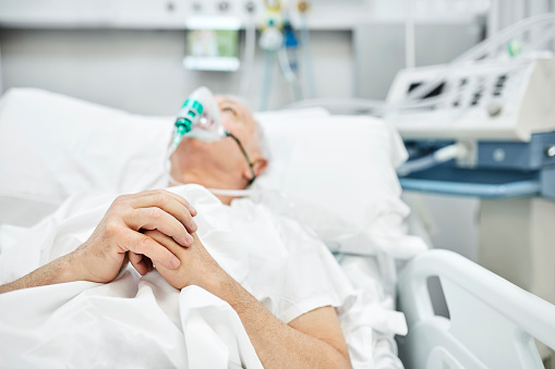 Senior man praying in ward. Male patient suffering from coronavirus is lying on stretcher. He is in hospital during pandemic.