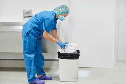 Side view of nurse disposing used protective workwear in garbage can. Female healthcare worker is working during coronavirus outbreak. She is in uniform at hospital.
